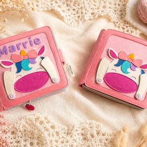 May include: Two pink felt books with unicorn designs. Each book features a unicorn face with a pink muzzle, white ears, and a colourful bow. One book has the name "Marrie" in purple letters. The books are on a cream-coloured surface.
