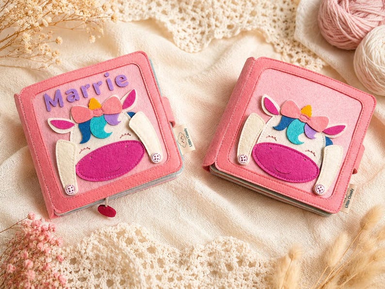 May include: Two pink felt books with unicorn designs. Each book features a unicorn face with a pink muzzle, white ears, and a colourful bow. One book has the name "Marrie" in purple letters. The books are on a cream-coloured surface.