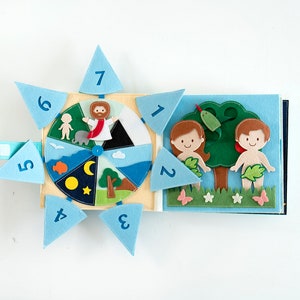 May include: A handmade felt book with a religious theme. The open book shows scenes from the Bible, including Adam and Eve, and a wheel depicting the days of creation. The book is made of various colors of felt.