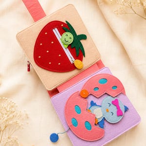 May include: A colourful felt activity book for babies, featuring a red strawberry with a zip and a baby bird hatching from an egg. The book is made of soft felt in shades of pink, red, beige, and blue, with a handle.