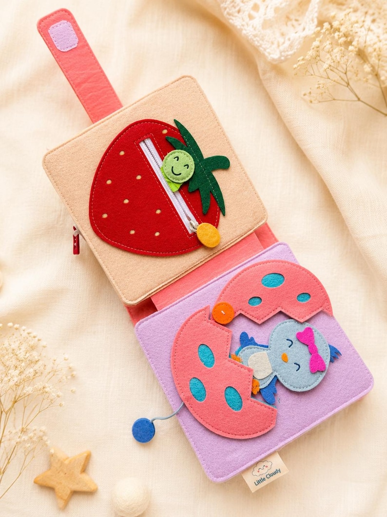 May include: A colourful felt activity book for babies, featuring a red strawberry with a zip and a baby bird hatching from an egg. The book is made of soft felt in shades of pink, red, beige, and blue, with a handle.