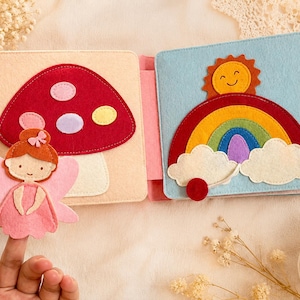 May include: A handmade felt book with colourful illustrations. One page shows a red mushroom with pink, purple, and yellow spots, and a pink fairy. The other page features a rainbow, smiling sun, and clouds against a blue background. The book is designed for children.