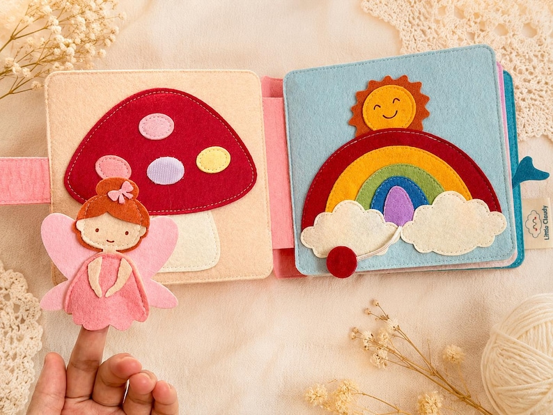 May include: A handmade felt book with colourful illustrations. One page shows a red mushroom with pink, purple, and yellow spots, and a pink fairy. The other page features a rainbow, smiling sun, and clouds against a blue background. The book is designed for children.