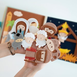 May include: A felt nativity scene with figures of Mary, Joseph, baby Jesus, an angel, sheep, a donkey, and a cow. The figures are held in front of an open felt book with illustrations of a stable and desert scene.
