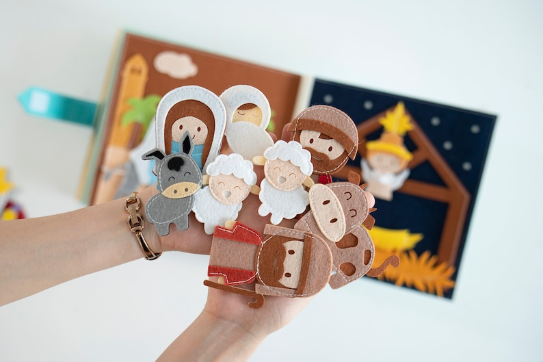 May include: A felt nativity scene with figures of Mary, Joseph, baby Jesus, an angel, sheep, a donkey, and a cow. The figures are held in front of an open felt book with illustrations of a stable and desert scene.