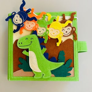 May include: A green felt dinosaur with a smiling face is surrounded by five felt monkeys hanging from a string. The monkeys are different colors: blue, orange, green, yellow, and brown. The background is brown felt with green felt leaves.
