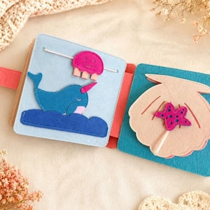 May include: A felt activity book featuring a narwhal, jellyfish, and seashell design. The pages are light blue and turquoise, with pink and blue accents. The book has a coral strap and is designed for children.
