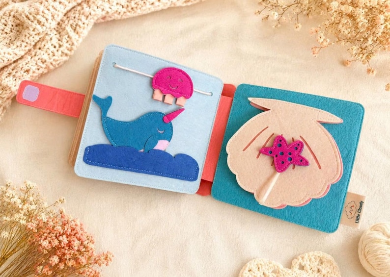 May include: A felt activity book featuring a narwhal, jellyfish, and seashell design. The pages are light blue and turquoise, with pink and blue accents. The book has a coral strap and is designed for children.