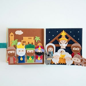 May include: A felt nativity scene book with a teal ribbon closure. The open book displays a scene with figures of the three wise men, angels, and animals. The background features buildings, palm trees, and a starry night.
