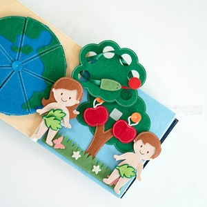 May include: A handmade felt quiet book with a biblical theme. The book features a felt Earth, Adam and Eve, an apple tree with red apples, and a green snake. The pages are blue, beige, and green.