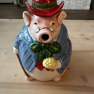 May include: A ceramic pig-shaped teapot wearing a red hat with a green band, a blue jacket with red stripes, and a green bow tie. The pig is holding a yellow flower and a cane. The teapot has a whimsical, vintage design.