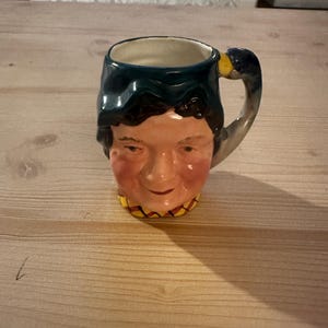 May include: A ceramic mug with a face design. The mug features a smiling face with rosy cheeks, dark blue hair, and a yellow and red collar. The handle is shaped like a duck's head. The mug is sitting on a wooden surface.