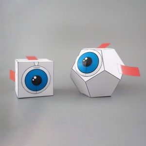 Pode incluir: Dois modelos de papel de um olho humano. O modelo à esquerda é um cubo com a palavra "córnea" impressa nele. O modelo à direita é uma forma 3D com um design de olho azul.