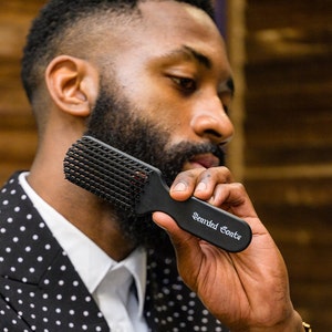May include: A man with a beard is holding a black beard brush with the words "Bearded Goats" printed on it. He is wearing a black and white polka dot blazer.