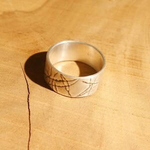 Op de afbeelding: Een zilveren ring met een getextureerd ontwerp. De ring is breed en heeft een gladde, gepolijste afwerking.