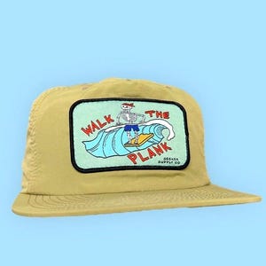 Puede incluir: Gorra de béisbol beige con un parche rectangular. El parche muestra un esqueleto surfeando una ola, con la frase "WALK THE PLANK". El fondo es azul claro. También se ve el texto "SEEKER SUPPLY CO".