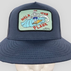 May include: A navy blue trucker hat with a black mesh back. The hat features a colourful patch with a skeleton surfing on a wave and the text "Walk the Plank" and "Seeker Supply Co."