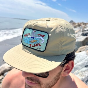 Puede incluir: Una gorra de béisbol beige con un parche que dice "Walk the Plank" y un esqueleto de dibujos animados haciendo surf. El parche es rojo, azul y blanco. El texto "Seeker Supply Co." está en el parche.
