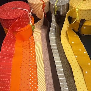 May include: A collection of fabric ribbon rolls in various colors and patterns, including red with white dashed lines, solid orange, orange with white polka dots, yellow and white diagonal stripes, brown, gray with white dashes, and yellow with white dots.