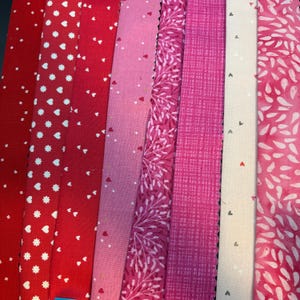 May include: A collection of red and pink fabric swatches. The fabrics feature various patterns, including white hearts, floral designs, and a grid pattern. The color palette ranges from deep red to light pink.