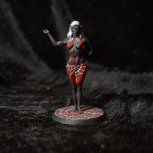 May include: Miniature figurine of a dark-skinned woman with white hair and pointed ears, wearing a red bodysuit with a spiderweb design. The figure is standing on a circular base with a detailed pattern, set against a dark background.