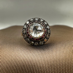 Può includere: Un anello rotondo con una pietra preziosa trasparente sfaccettata con una montatura in argento. L'anello è circondato da un alone di diamanti più piccoli e una fila di rubini rossi.