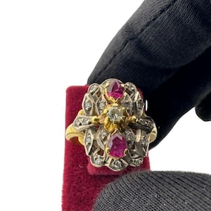 May include: An ornate gold ring featuring a central diamond and two pear-shaped rubies. The ring is further embellished with smaller diamonds and intricate filigree detailing, set against a red velvet background.