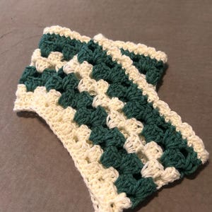 May include: A rectangular crochet piece featuring a repeating granny square pattern with alternating diagonal stripes of cream and dark forest green yarn. The textile has a textured, open-weave construction with visible yarn loops throughout.