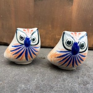 Op de afbeelding: Twee keramische uilenbeeldjes met een crèmekleurige basis en handgeschilderde details in blauw en oranje. Elke uil heeft een opvallende blauwe snavel, grote ronde ogen en een stralend patroon van blauwe en oranje lijnen op de borst.