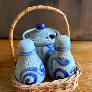 May include: A ceramic salt and pepper shaker set with a matching lidded condiment jar, all featuring a light blue speckled glaze with dark blue painted floral patterns. The shakers are marked with S and P, and the set sits in a woven wicker basket.