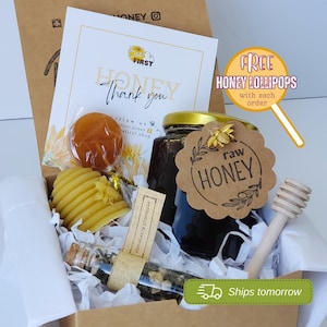 May include: A gift box filled with honey-themed items, including a jar of raw honey, a honeycomb-shaped candle, a small jar of honey-infused tea, a honey lollipop, and a wooden honey dipper. The box is decorated with a tag that says "Free Honey Lollipops with each order".