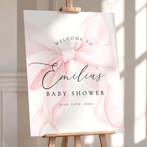 May include: A rectangular welcome sign for a baby shower featuring a large, soft pink watercolor bow illustration. The text reads: Welcome to Emilia's Baby Shower, June 24th, 20XX. The sign is displayed on a wooden easel stand against a neutral wall.