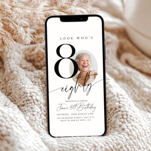 Puede incluir: Un smartphone muestra una invitación de cumpleaños. El diseño incluye el texto "LOOK WHO'S 80 eighty" y una foto de una mujer sonriente. También hay detalles del evento. El teléfono está colocado sobre una superficie texturizada de color crema.
