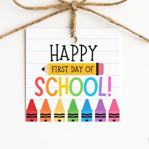 Könnte beinhalten: Ein quadratisches weißes Geschenkanhänger-Schild mit blauen horizontalen Linien, die an Notizbuchpapier erinnern. Der Text lautet HAPPY FIRST DAY OF SCHOOL mit einem gelben Bleistift. Sieben bunte Wachsmalstifte in Rot, Orange, Gelb, Grün, Blau, Lila und Pink.