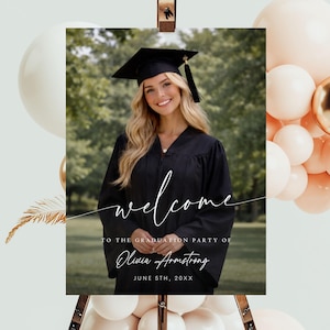 May include: A graduation welcome sign featuring a smiling woman in a cap and gown. The sign includes the word "welcome" in elegant script, the graduate's name, and the date. The sign is displayed on an easel with balloons in the background.