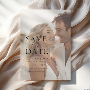 May include: A wedding save-the-date card with a photograph of a smiling couple. The card includes the text "SAVE THE DATE" in a stylish script, the names Olivia & Jameson, the wedding date, and the location. The background is a soft, neutral colour.
