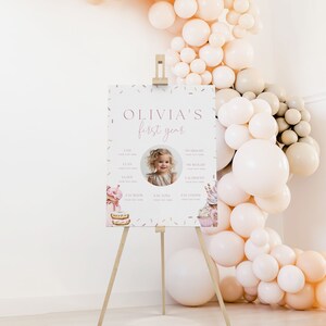 May include: A pink and white printable first birthday sign with a photo of a girl, confetti, and a sweet theme. The sign includes the text "Olivia's first year" and sections for the child's age, height, weight, and favorite things.