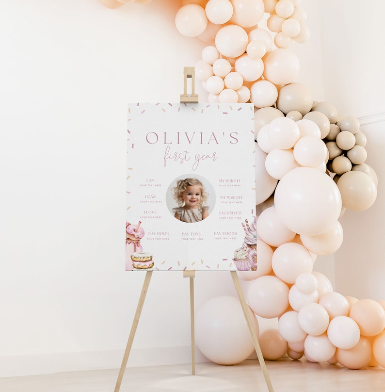 May include: A pink and white printable first birthday sign with a photo of a girl, confetti, and a sweet theme. The sign includes the text "Olivia's first year" and sections for the child's age, height, weight, and favorite things.