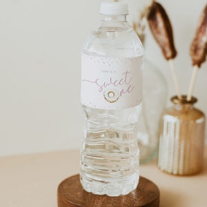May include: A clear plastic water bottle with a white label that says "She's a sweet one" with a pink donut graphic. The bottle is on a wooden coaster.