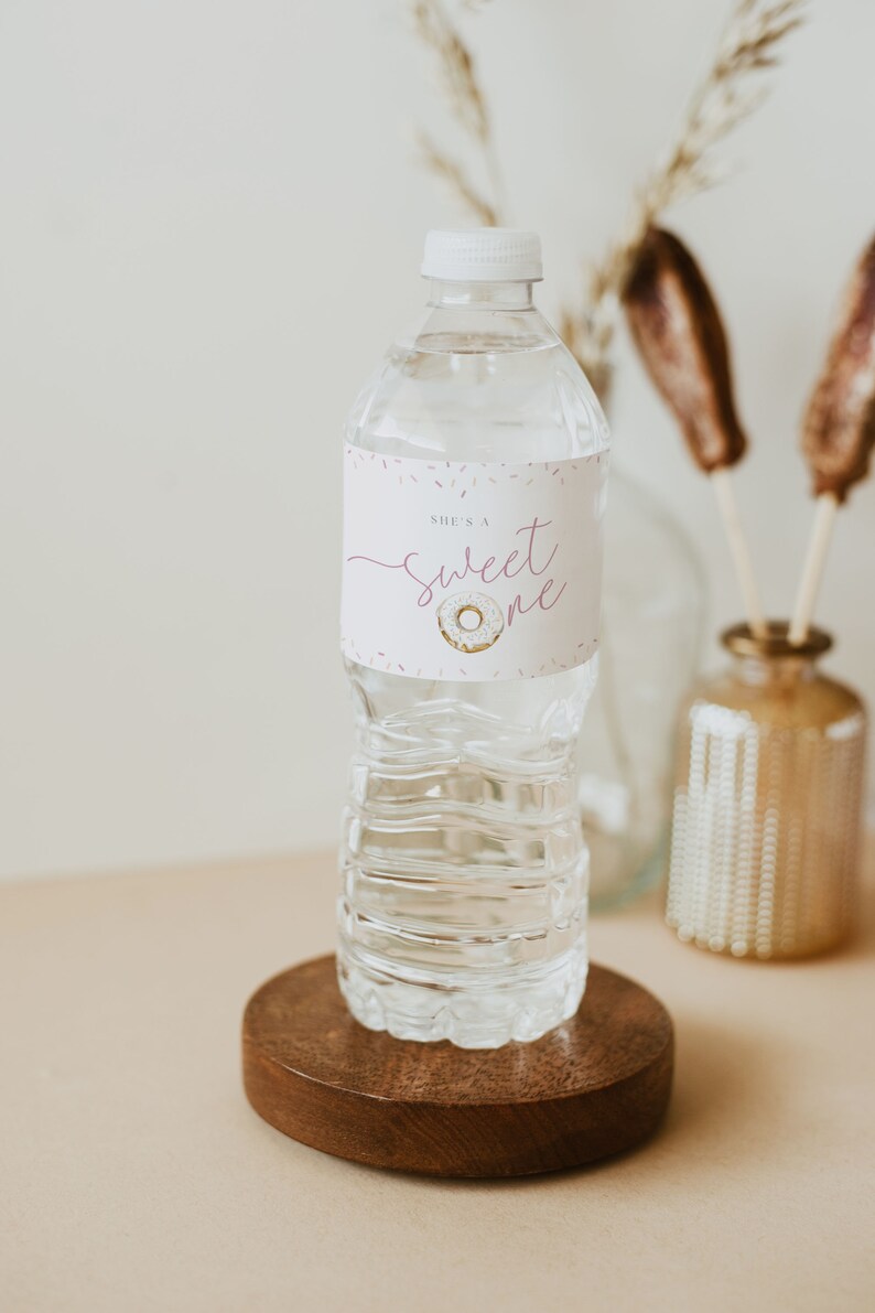 May include: A clear plastic water bottle with a white label that says "She's a sweet one" with a pink donut graphic. The bottle is on a wooden coaster.