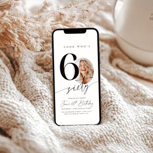 May include: A smartphone displays a birthday invitation with the text "Look Who's 60". The design features a photo of a smiling woman, large black numbers, and elegant script. The phone rests on a soft, textured surface.