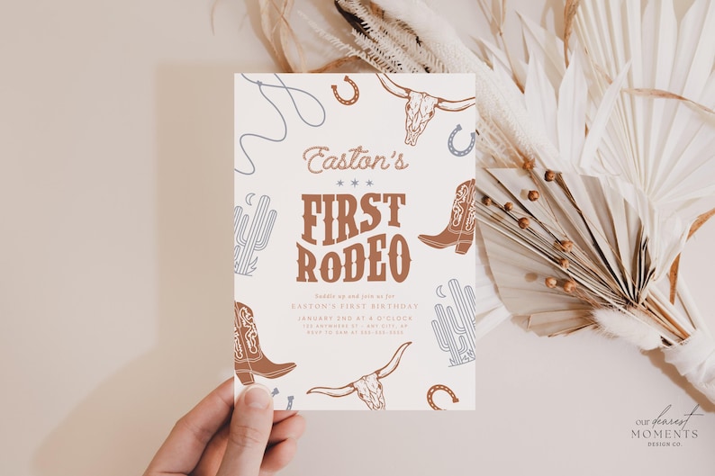 May include: A rectangular invitation card with a western theme. The card features the text "Easton's First Rodeo" in brown and white, along with illustrations of cowboy boots, a bull skull, and a horseshoe. The card is held by a hand.