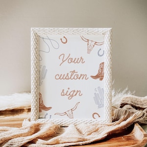 May include: White wooden frame with a white background and a brown rope design that says "Your custom sign". The background is decorated with a pattern of cacti, cowboy boots, horseshoes, and bull skulls.