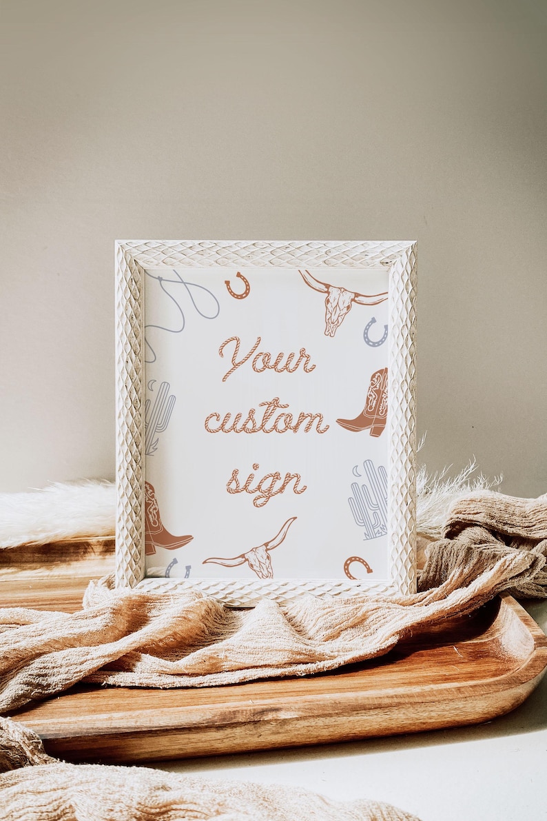 May include: White wooden frame with a white background and a brown rope design that says "Your custom sign". The background is decorated with a pattern of cacti, cowboy boots, horseshoes, and bull skulls.