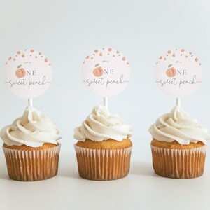 May include: Three cupcakes with white frosting topped with round peach-themed cake toppers that say "One sweet peach".