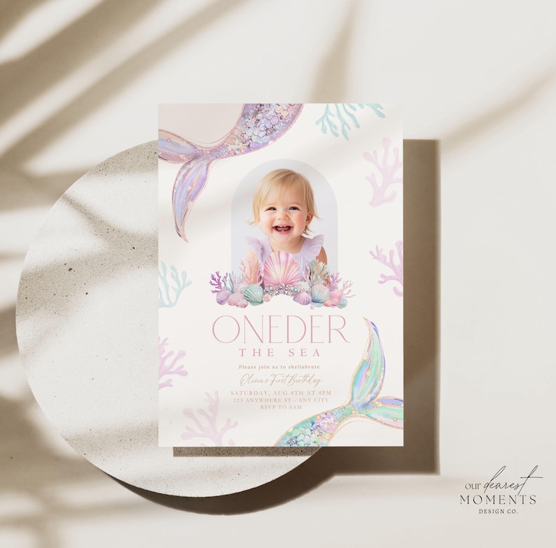 May include: A pink and blue mermaid-themed first birthday invitation with a photo of a baby girl in a seashell crown. The invitation says "ONEDER THE SEA" and includes the date and time of the party.