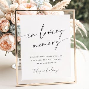 May include: A framed sign with gold trim and the words "in loving memory" in elegant script. The sign includes the phrases "Remembering those who are not here, but will always be in our hearts" and "today and always". Soft floral arrangements are in the background.