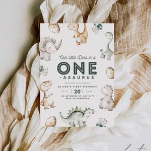May include: A white invitation card with a green and brown dinosaur illustration. The text reads "Our little Dino is a ONE -ASAURUS. Please join us to celebrate OLIVER'S FIRST BIRTHDAY. SEPT | 20 | 2PM. 123 ANYWHERE ST, ANY CITY. RSVP TO SAMANTHA. our dearest MOMENTS DESIGN CO."