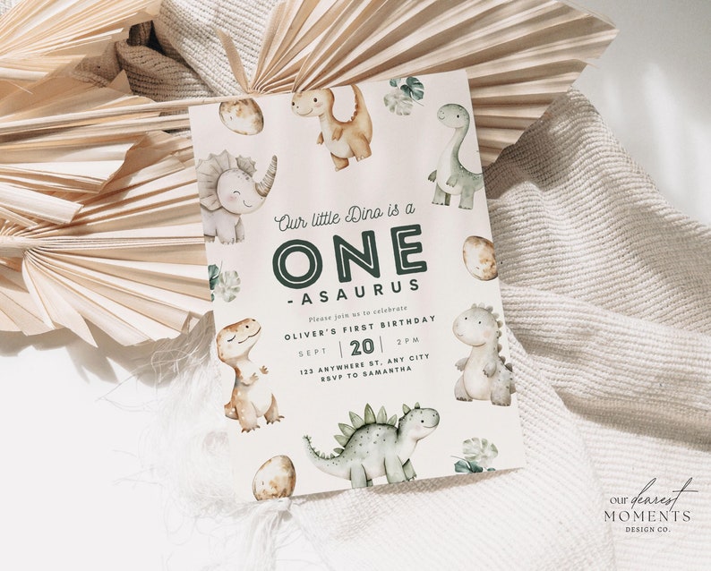 May include: A light pink invitation with green and brown dinosaur illustrations. The text reads "Our little Dino is a ONE -ASAURUS. Please join us to celebrate OLIVER'S FIRST BIRTHDAY SEPT 20 2PM 123 ANYWHERE ST. ANT CITY RSVP TO SAMANTHA".