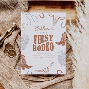 May include: A white invitation card with a rodeo theme. The text "Easton's First Rodeo" is displayed in brown, along with illustrations of cowboy boots, cacti, and horseshoes. The card is on a wooden tray with a beige fabric.